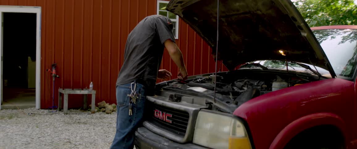 A man in jeans and a t-shirt works on the engine of his red GMC truck with the hood up in front of a red barn