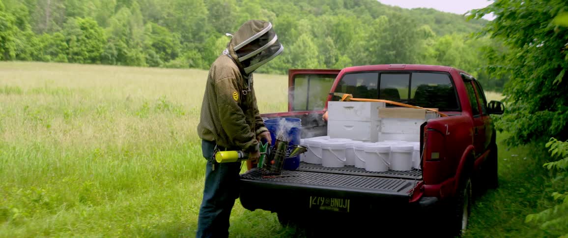 A beekeeper in a protective suit prepares a bee smoker on the tailgate of a truck in a rural field