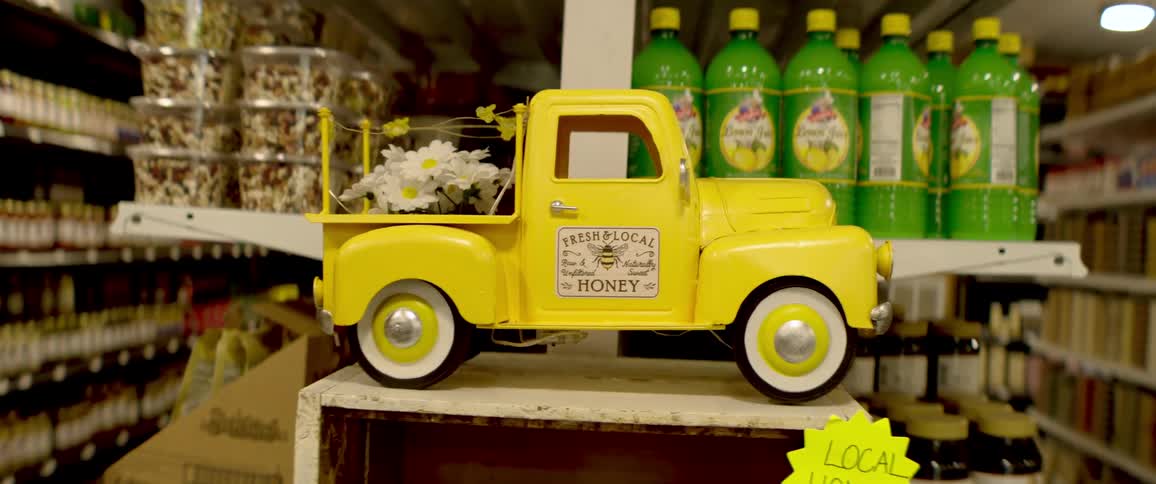 A charming yellow miniature truck with daisies in the back advertises fresh and local honey in a country store