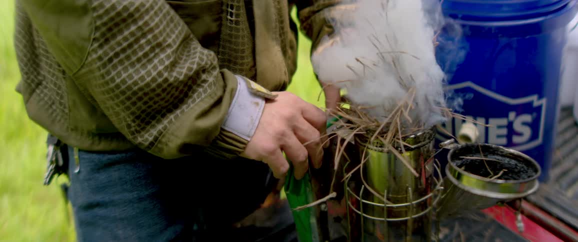 Slow motion close-up shot of a beekeeper lighting a bee smoker with pine needles before tending to the hives