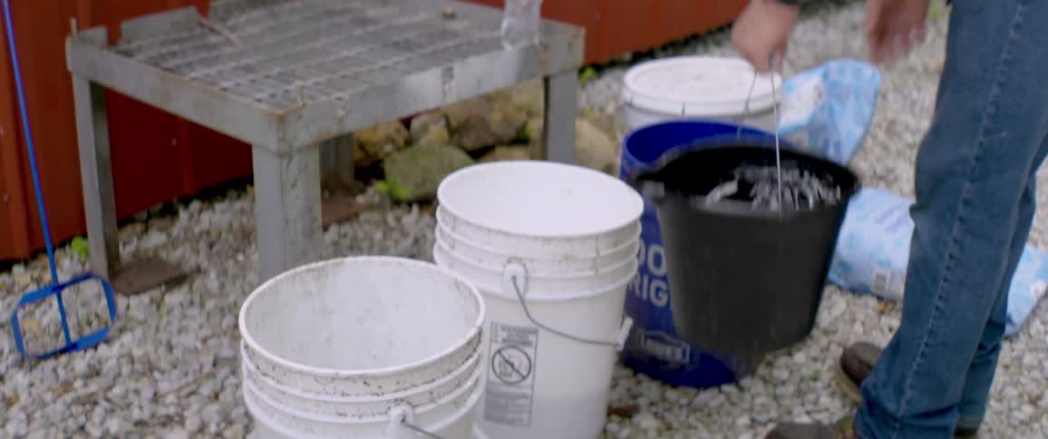 A man in jeans pours granulated sugar from a bag into a bucket, then adds water to create a mixture outdoors