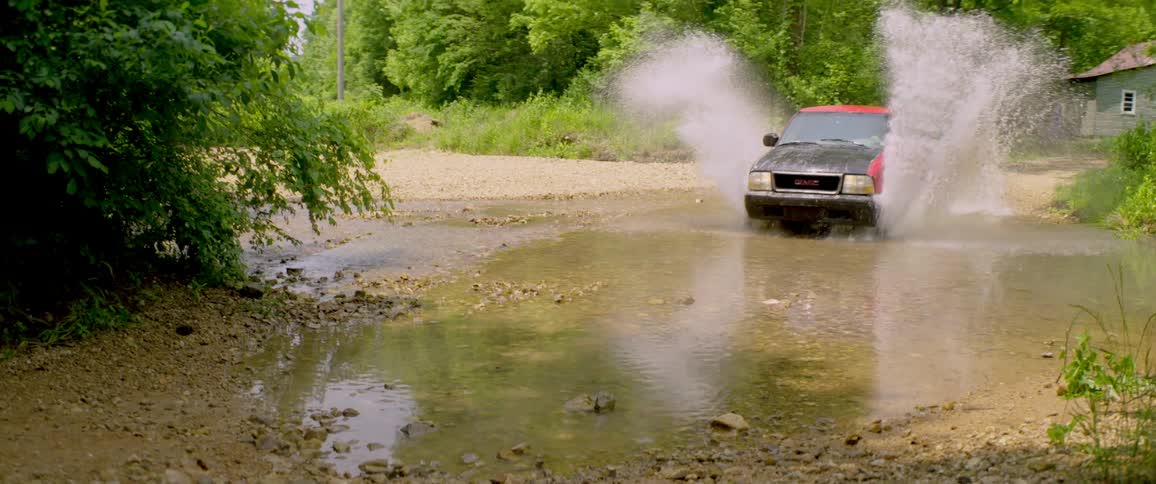 A red and black pickup truck splashes through a shallow stream in the countryside in slow motion