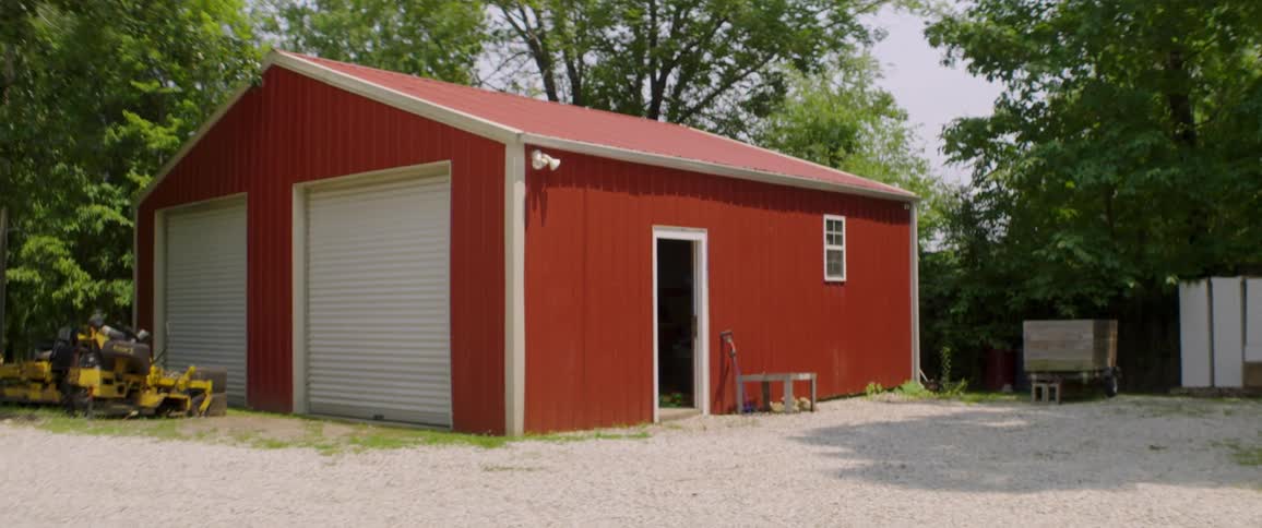 A static shot of a red metal garage with white roll-up doors on a gravel lot surrounded by trees