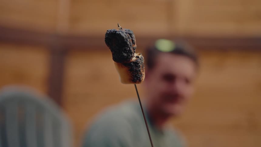 A close-up shot of a burnt marshmallow held up on a roasting stick with a man out of focus in the background