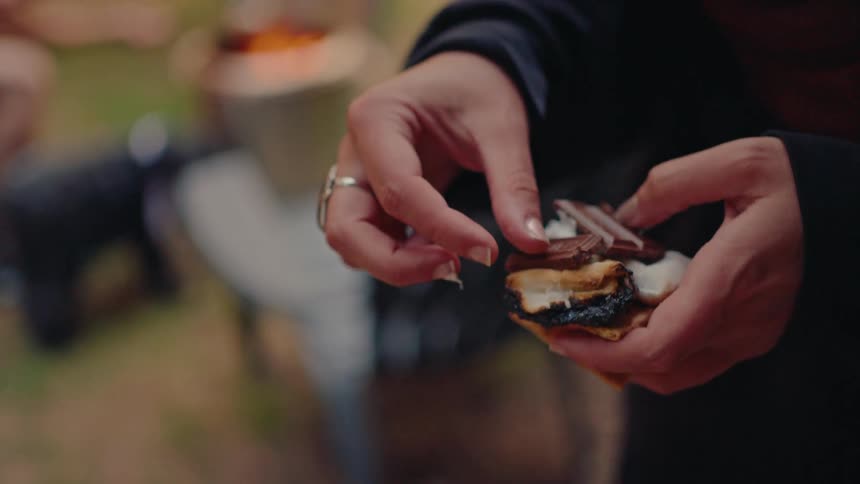A person carefully assembles a gooey, toasted s'more with chocolate in slow motion next to a backyard fire pit