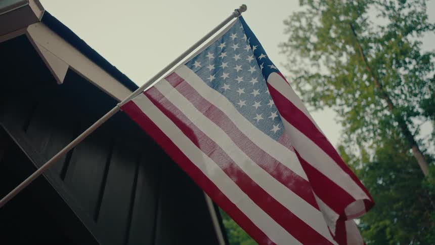 From a low angle, the American flag waves gently in the breeze while mounted on the side of a rustic home