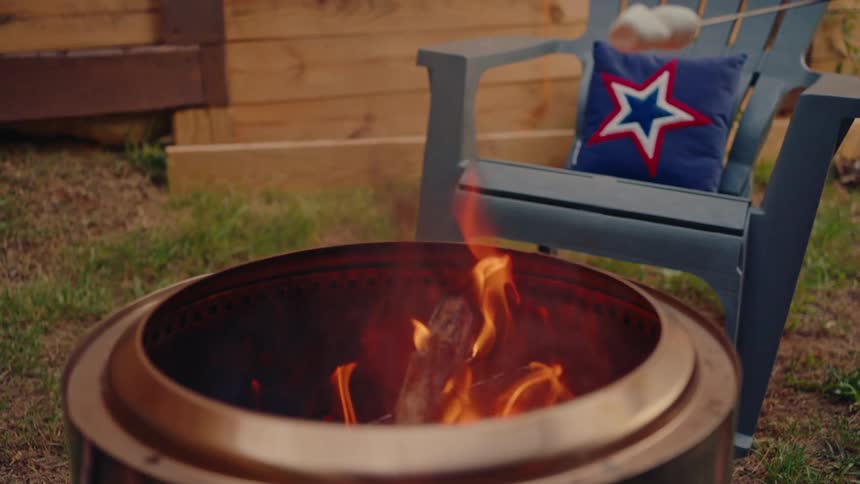 Roasting marshmallows in slow motion over a backyard fire pit with a patriotic star pillow on a chair in the background