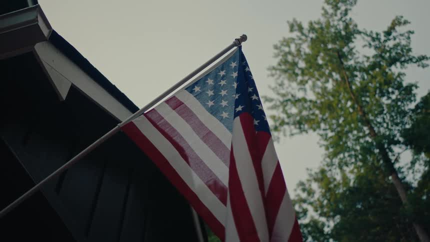 A low-angle static shot of an American flag hanging from a house on an overcast day with trees in the background