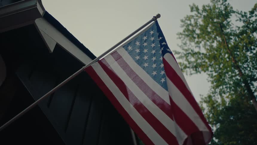 A low angle shot of a still American flag hanging from the side of a house with trees in the background