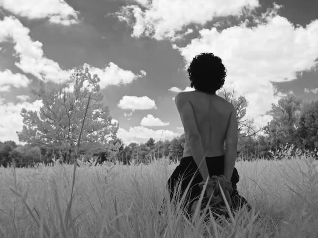 A shirtless person with hands tied behind their back slowly kneels in a grassy field in this dramatic black and white clip