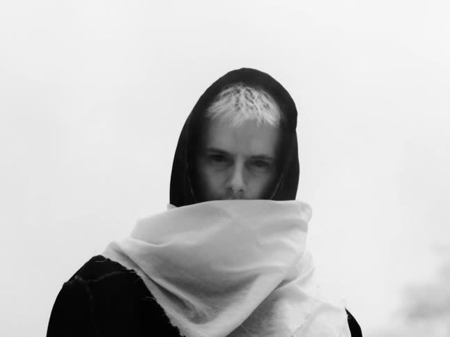 A mysterious figure with striking white hair, cloaked in a dark hood and scarf, fixes an intense gaze on the viewer in this minimalist black and white clip