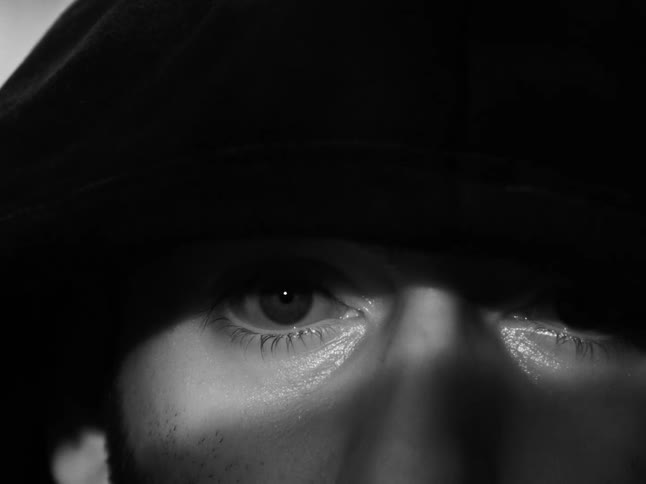 The intense, focused eyes of a mysterious hooded figure emerge from the darkness in a black and white close-up