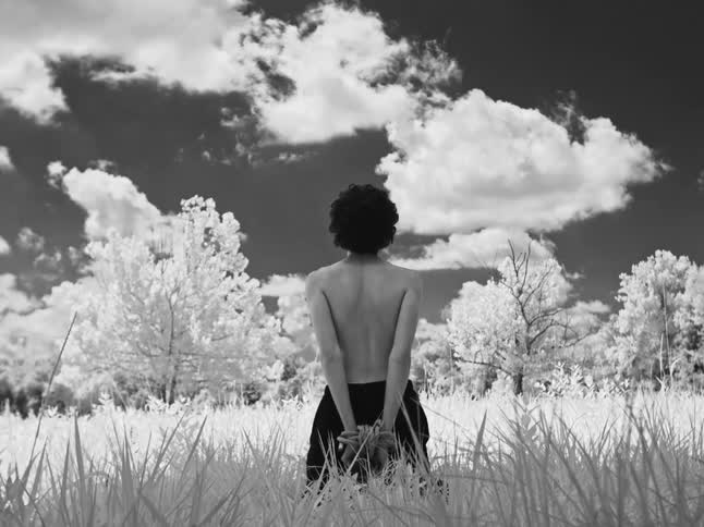 Artistic black and white still shot of a shirtless person with hands bound, standing in a surreal, dreamlike field
