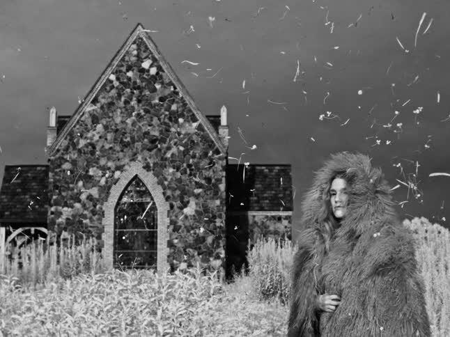 A surreal black and white cinemagraph shows a woman in a fur coat standing stoically before a stone church as particles drift by