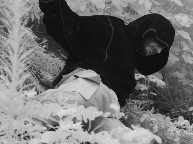 Infrared black and white shot of a man in a dark hoodie standing among surreal, glowing white foliage in a forest