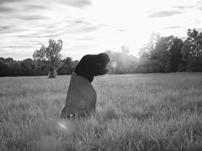 A mysterious figure in a dark hoodie and skirt dances slowly and expressively in a sunlit grassy meadow in black and white