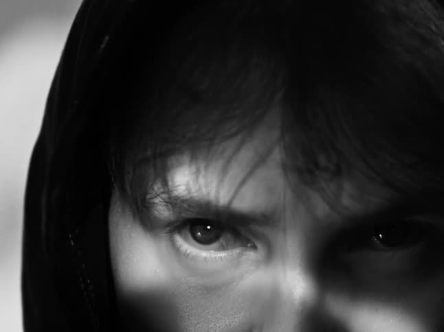 A close-up of a hooded figure slowly looking up with an angry and intense stare in black and white