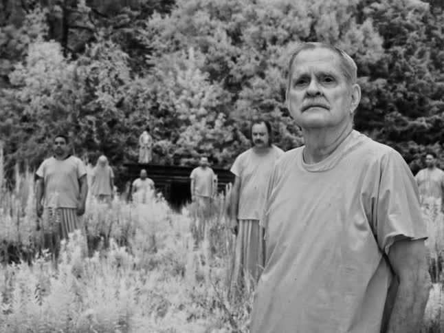 An eerie black and white still shot of a group of men standing motionless in a surreal, dreamlike forest