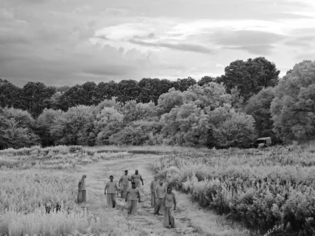 Somber black and white infrared footage of a group of men walking in a solemn procession through a surreal, overgrown field