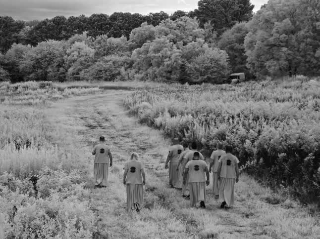 A surreal black and white infrared view shows a group of people in uniform walking down a rural path as lightning flashes in the sky
