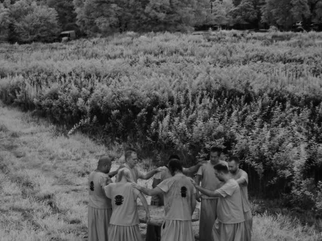 Black and white footage of a group of men standing in a huddle in a large, overgrown field