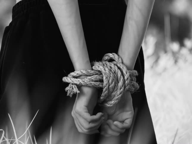 A somber black and white cinemagraph shows a person standing still with their hands bound by a thick rope behind their back