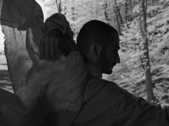 A dramatic black and white slow-motion shot of a man in a hooded robe as fabric billows slowly over his shoulder in a forest