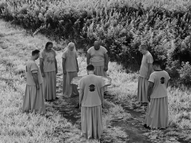 A static, high-angle, black and white shot shows a group of robed men standing in a mysterious circle in a wild, grassy field