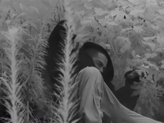 A mysterious man in a dark hood slowly awakens and sits up in an ethereal, dreamlike forest shot in surreal infrared