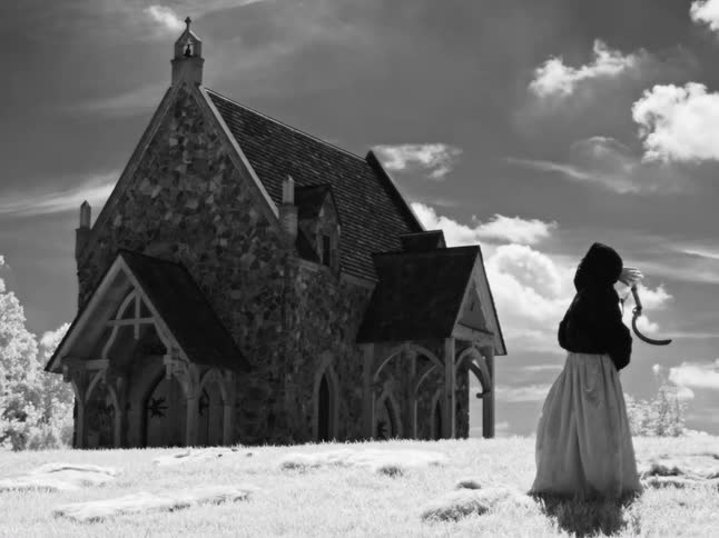 A surreal black and white cinemagraph shows a cloaked female figure resembling the Grim Reaper holding a scythe and looking at an old stone church