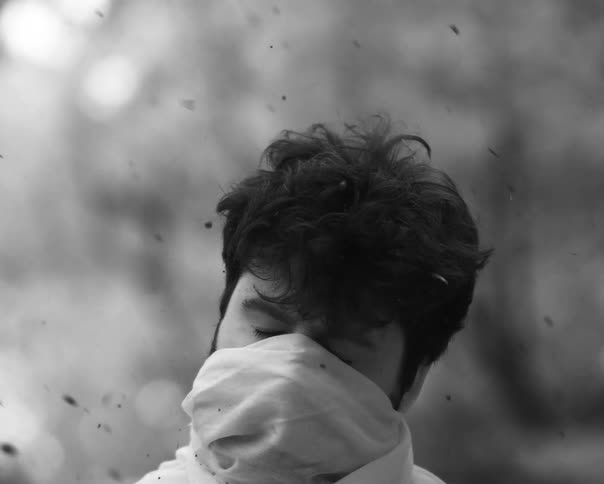 A man covers his face with a scarf as ash falls around him in a black and white slow-motion shot
