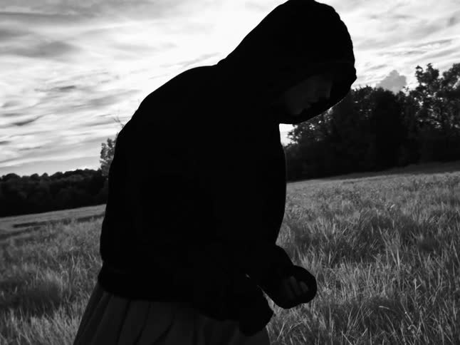 Dramatic black and white slow-motion shot of a lonely, hooded young man walking through a sunlit field