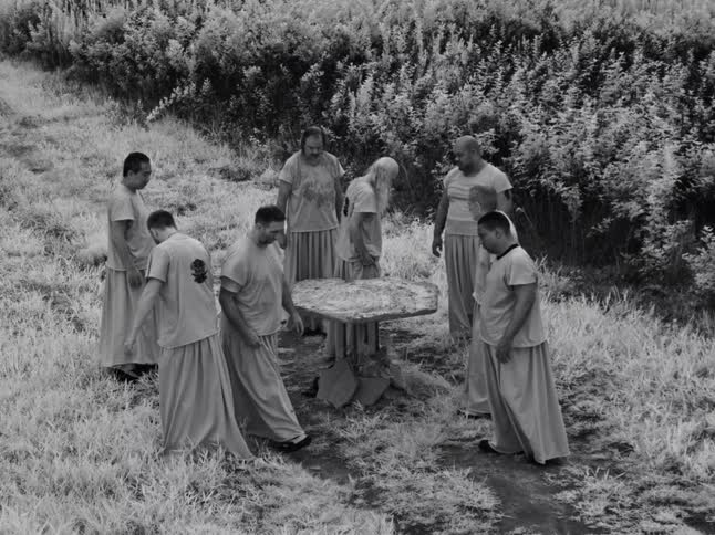 An eerie high-angle, black and white shot shows a strange gathering of men in robes standing motionless around a table in a field