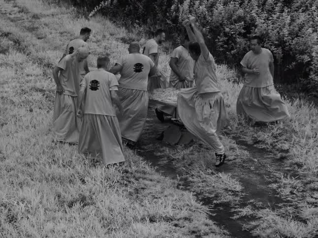 A group of men in uniform perform a surreal ritualistic practice in a grassy field, with one man seemingly levitating in a martial arts pose in this black and white, slow-motion clip