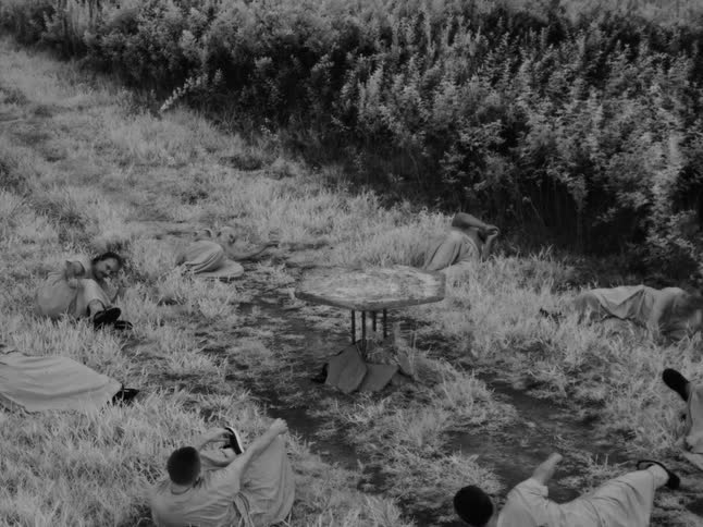 A surreal, black and white scene of robed figures slowly waking up in a grassy field around a mysterious table