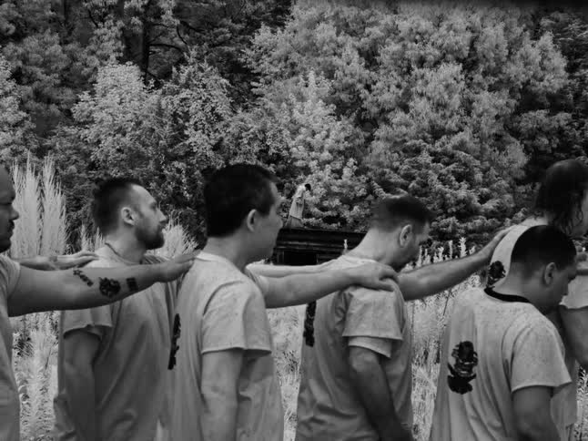 A slow-motion, black and white depiction of men walking in a single-file line through a field, each with a hand on the next person's shoulder