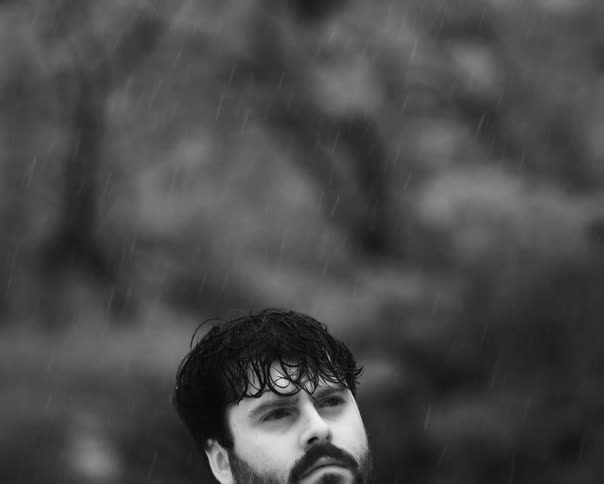 Black and white slow-motion shot of a dejected man standing in the rain as he slowly looks down