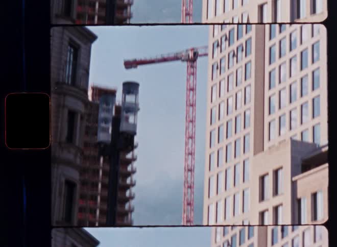 An external construction elevator rises alongside a building in the city with a red crane in the background on a vintage film