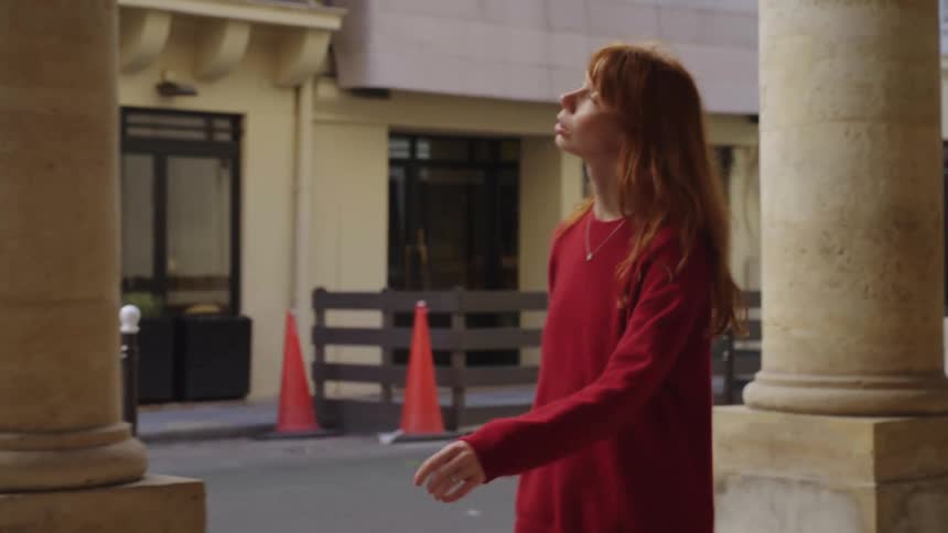 A young woman with long red hair walks in slow motion through a classic stone colonnade in the city