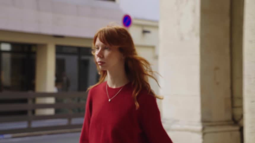 A stylish young woman with red hair walks in slow motion through a beautiful European colonnade on a breezy day