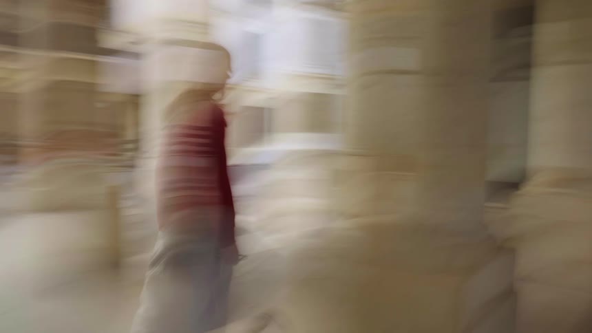 A disorienting slow-shutter shot follows a woman in a red sweater as she walks through a surreal, blurry colonnade