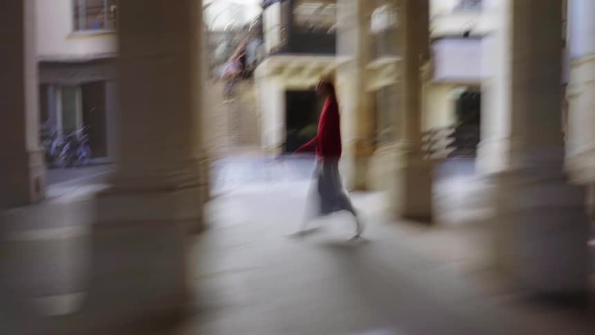 An impressionistic, motion-blurred side-view of a woman in red walking through a European colonnade