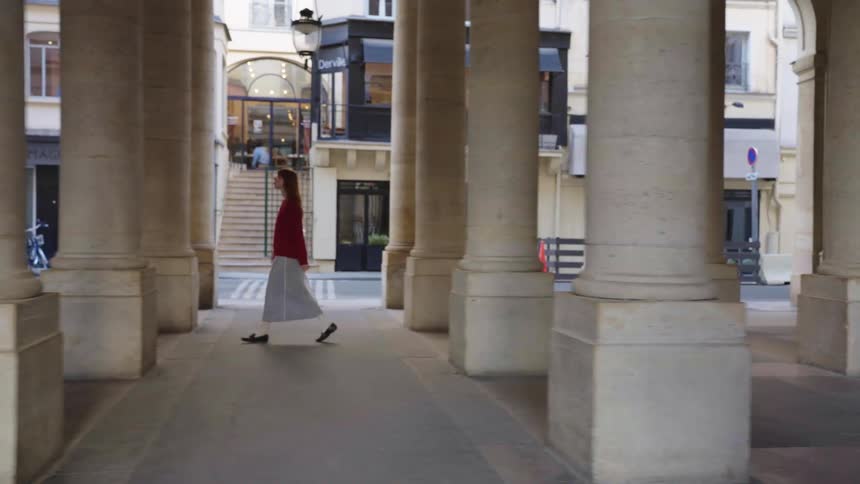 A slow-motion side view captures a stylish young woman walking through an elegant architectural colonnade in a European city