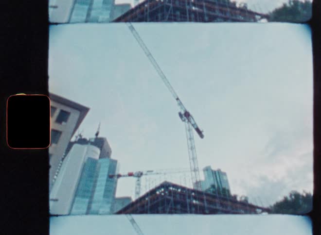 A vintage, low-angle film shot of a bustling city construction site with tall skyscrapers and cranes under a gray sky