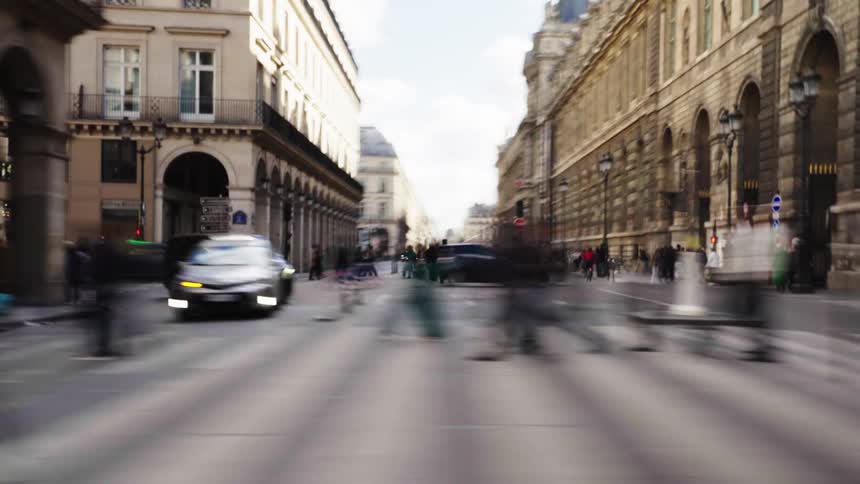 An artistic time-lapse shows blurred pedestrians and traffic moving through a busy street in Paris, France