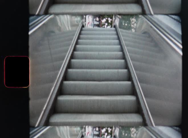 A low angle point-of-view of an escalator ascending, captured on vintage Super 8 film with a repetitive, abstract effect