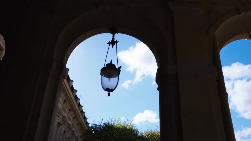 Vintage lanterns hang within a silhouetted archway, framing a view of a bright blue sky and a historic building