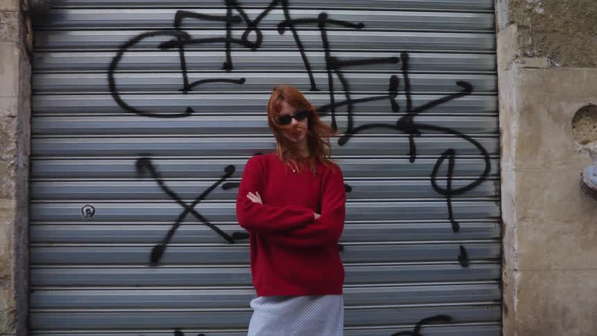 A stylish redhead woman with sunglasses poses in front of a graffiti-covered wall as the camera slowly zooms in