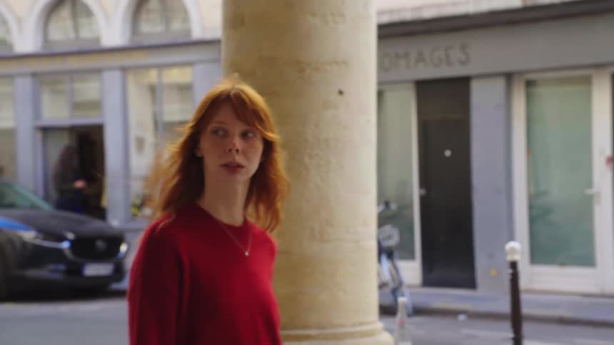 A pensive young woman with vibrant red hair walks in slow motion along a classic European street with large stone columns