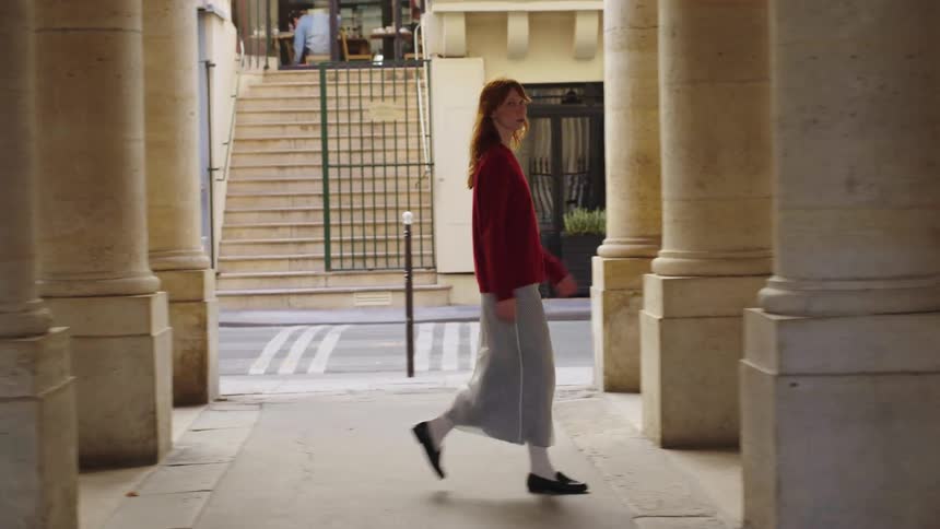 A stylish young woman with long red hair walks in slow motion along an outdoor colonnade